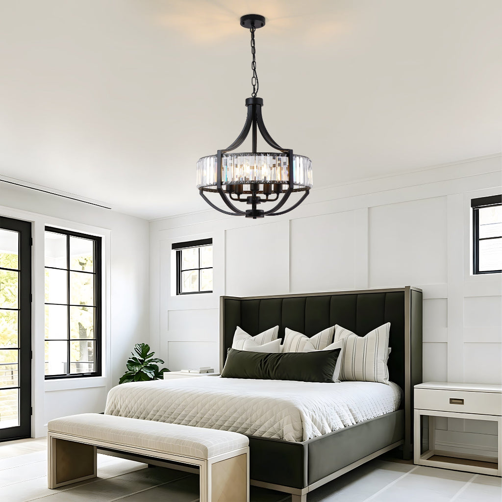 A modern bedroom interior featuring a black metal multi-light chandelier with clear cylindrical glass shades hanging from the ceiling. The room includes a bed with white bedding, a matching bench, and white paneled walls.
