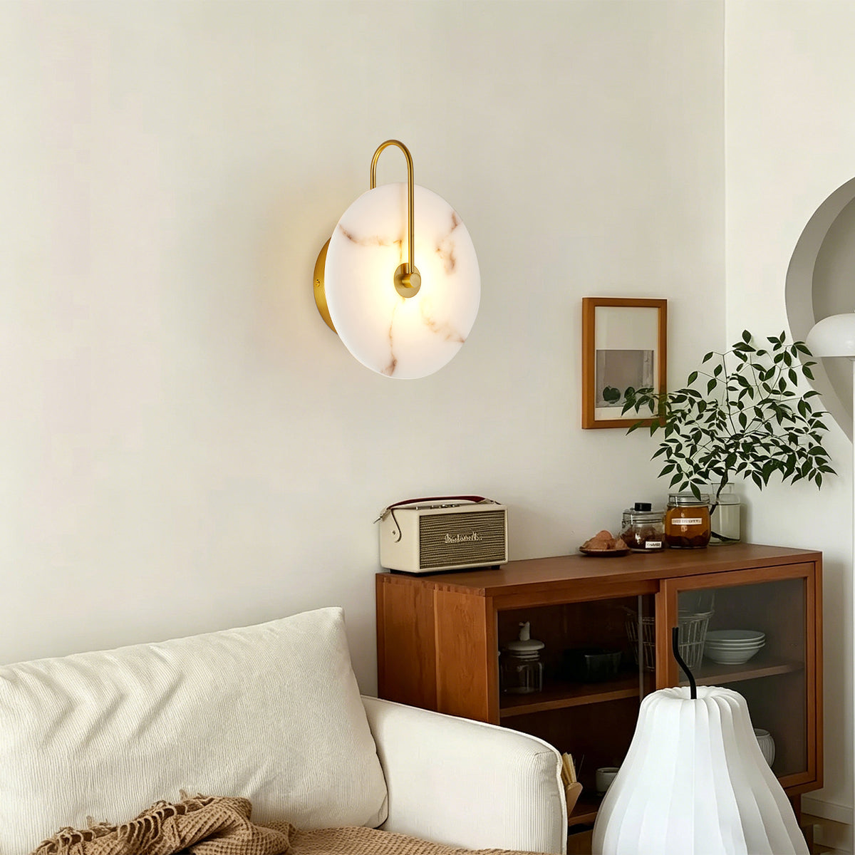A modern wall-mounted sconce with a round white marble-look shade and curved gold metal arms, glowing warmly above a wooden sideboard in a styled living room.