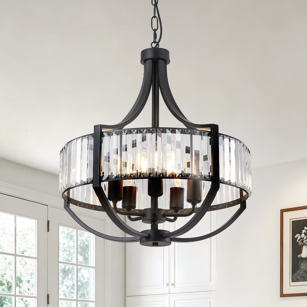 A round black metal chandelier with clear glass cylinder prisms and visible warm light bulbs, hanging in a bright white room with windows and framed wall art.