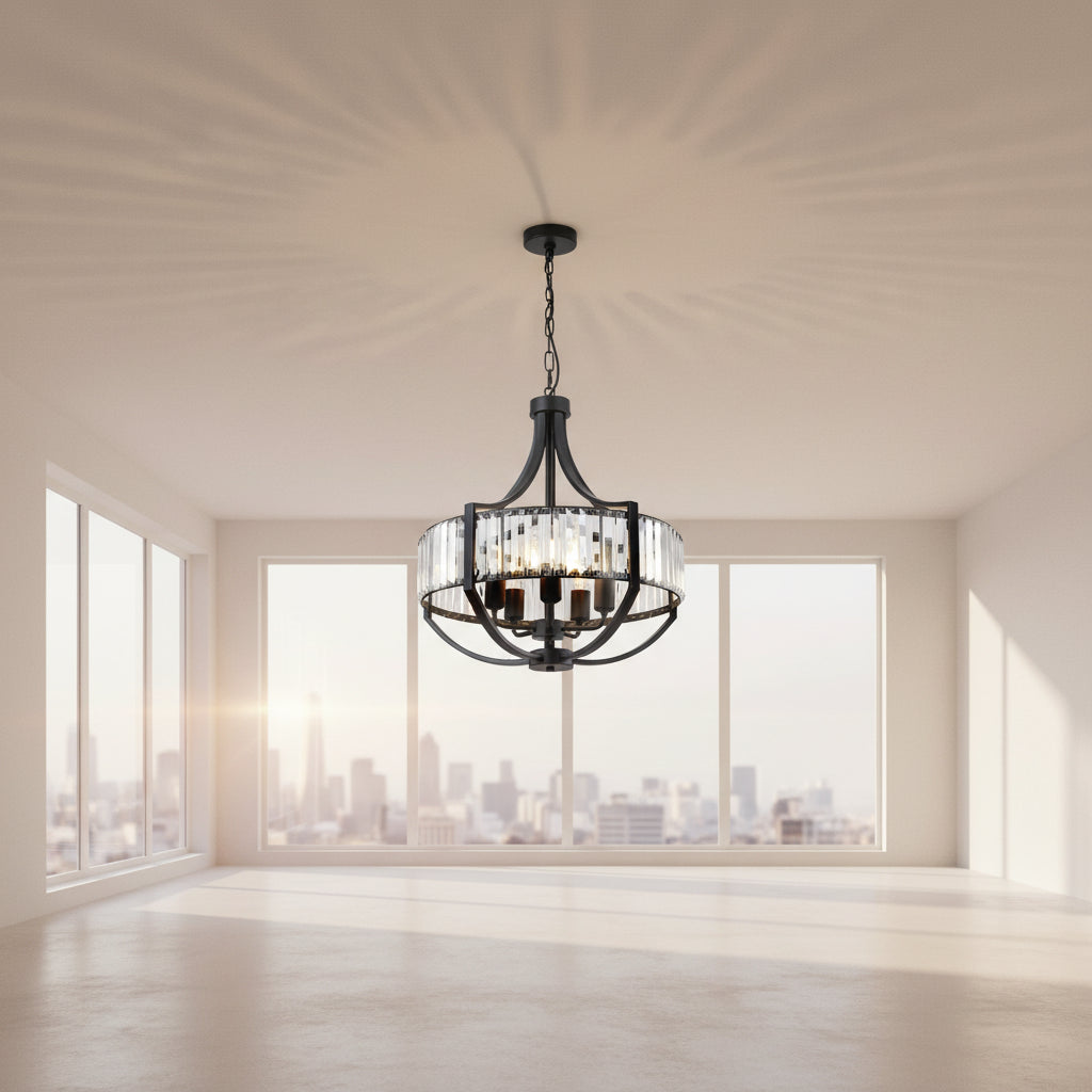 A round black metal chandelier with clear glass cylinder prisms and visible warm light bulbs, hanging in a bright white room with windows and framed wall art.